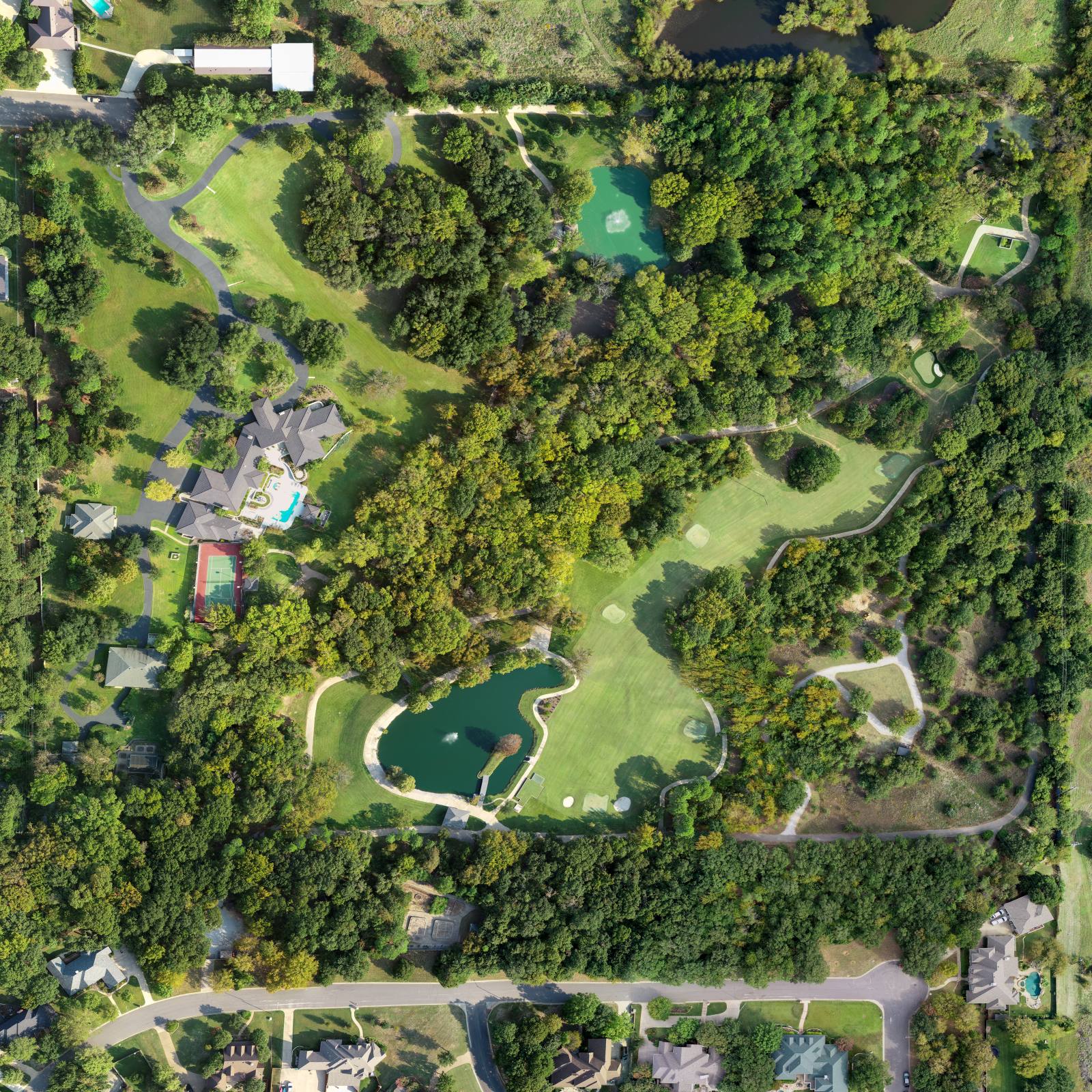 Aerial view of a golf course with trees, water hazards, and surrounding residential homes, including a large house with pool and tennis court.