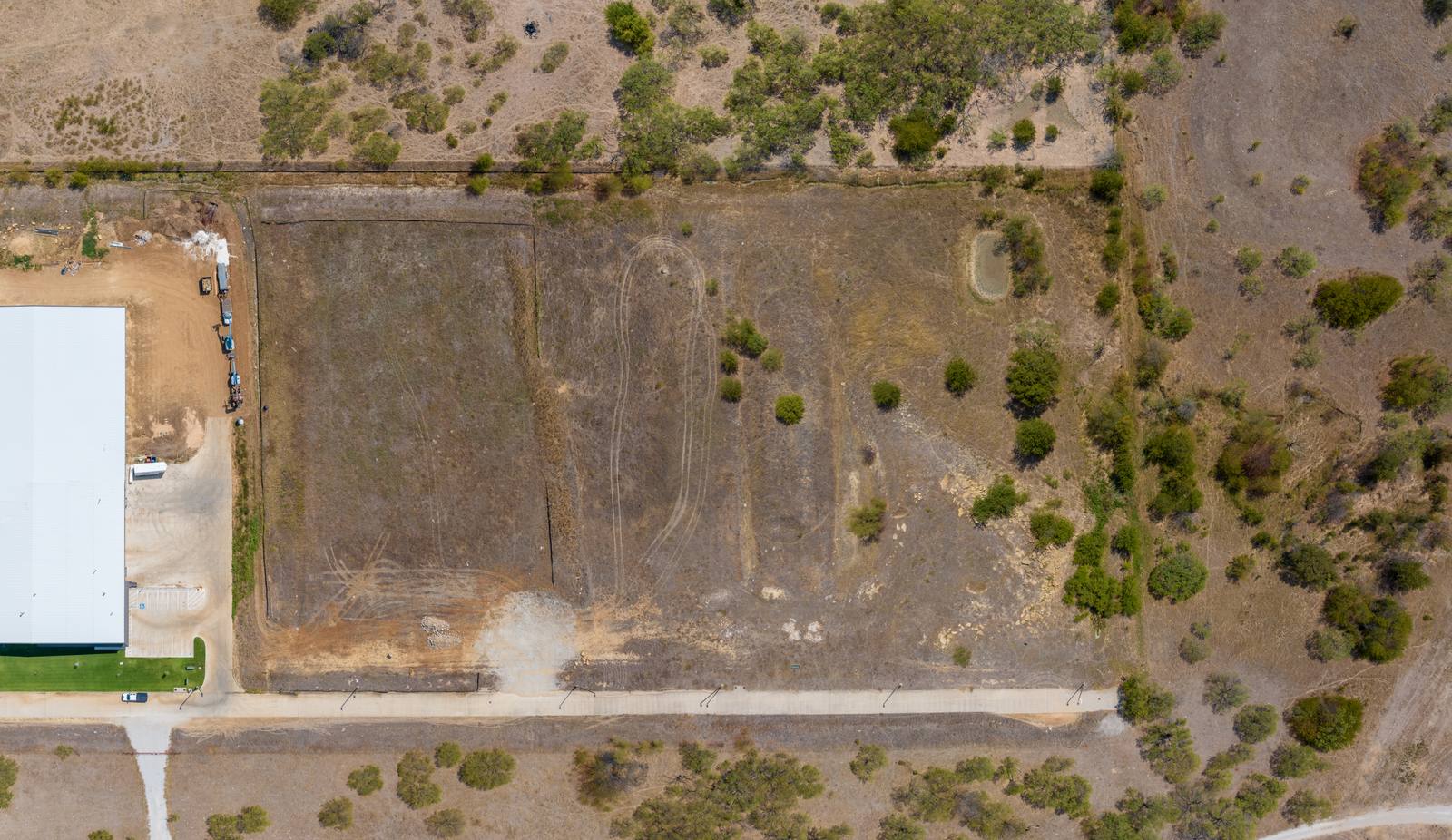 Aerial top-down view of a rectangular cleared lot bordered by sparse vegetation, a white building, and a road, with visible tire tracks and scattered debris.