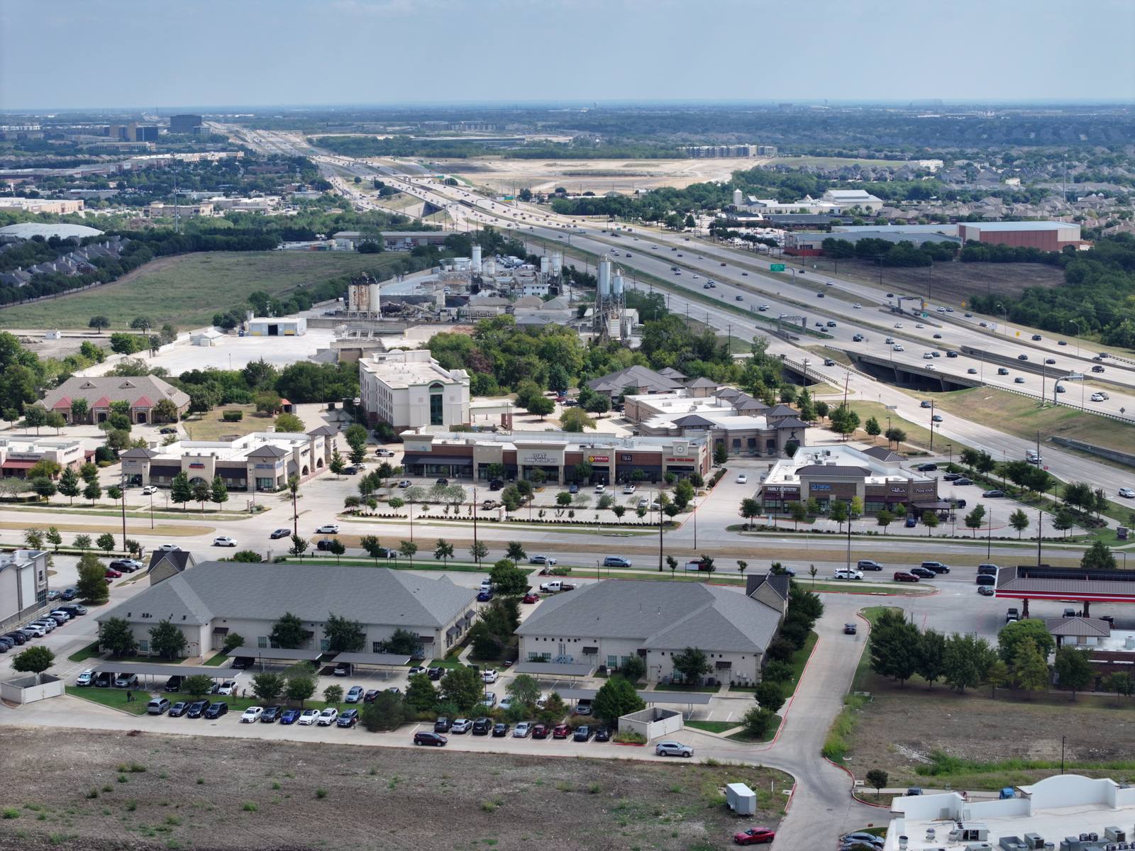 Aerial view of suburban commercial area with parking lots, buildings, and a multi-lane highway with moving vehicles under an overcast sky.