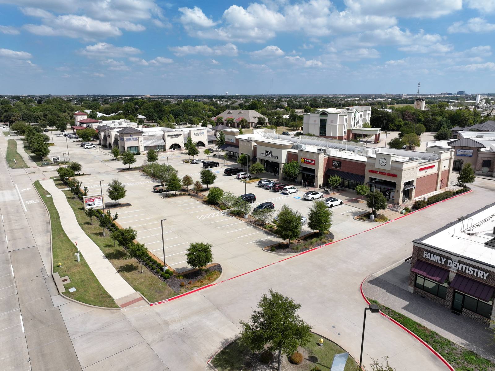 Aerial view of a commercial plaza with multiple storefronts, parking lots, and landscaped areas under a partly cloudy sky. No people visible.