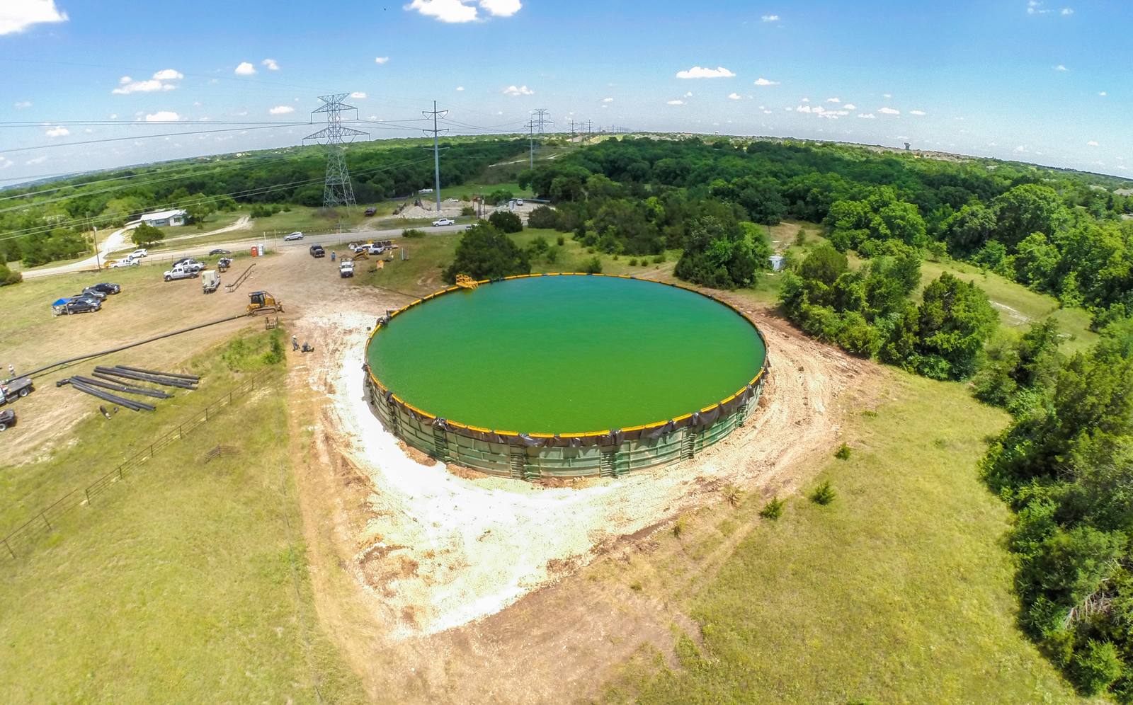 Aerial view of a large, circular, green-filled tank under construction in a grassy field, surrounded by trees, parked vehicles, and power lines under a partly cloudy sky.