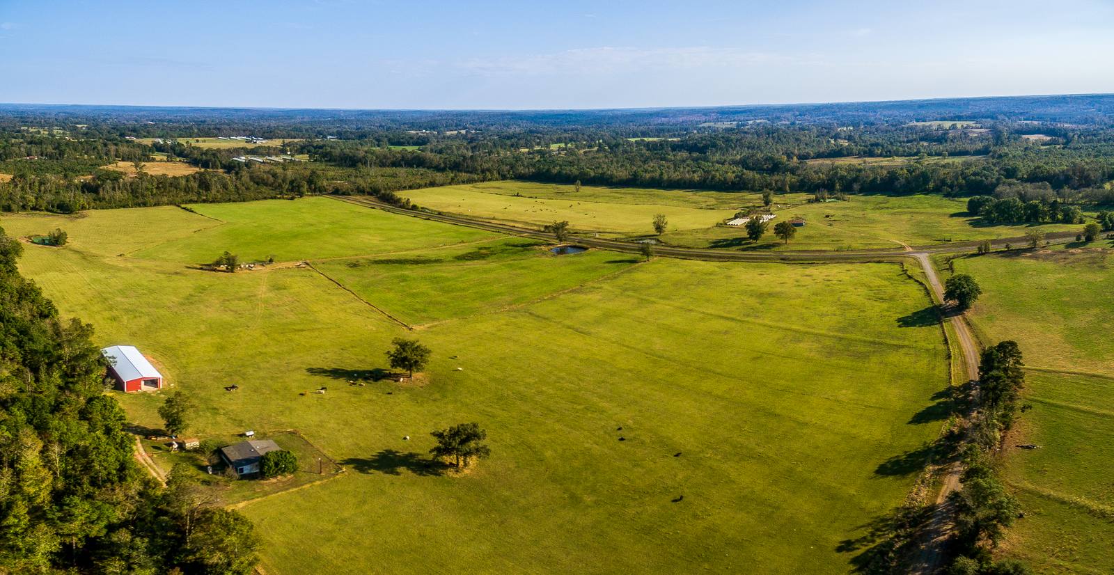 Aerial view of a rural landscape with green pastures, scattered trees, and a red-roofed barn, showing distant treelines under a clear sky.