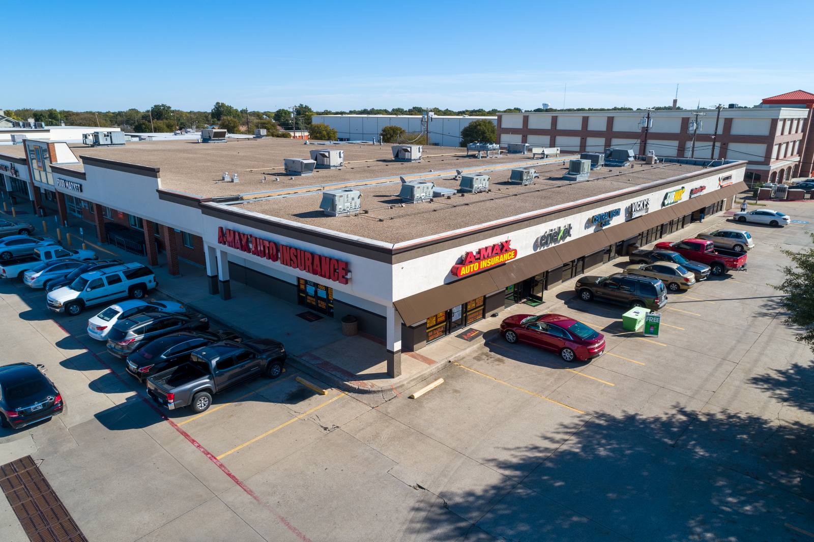 Aerial view of a commercial building with "A-MAX AUTO INSURANCE" signage, multiple parked cars, and rooftop HVAC units under clear blue sky.