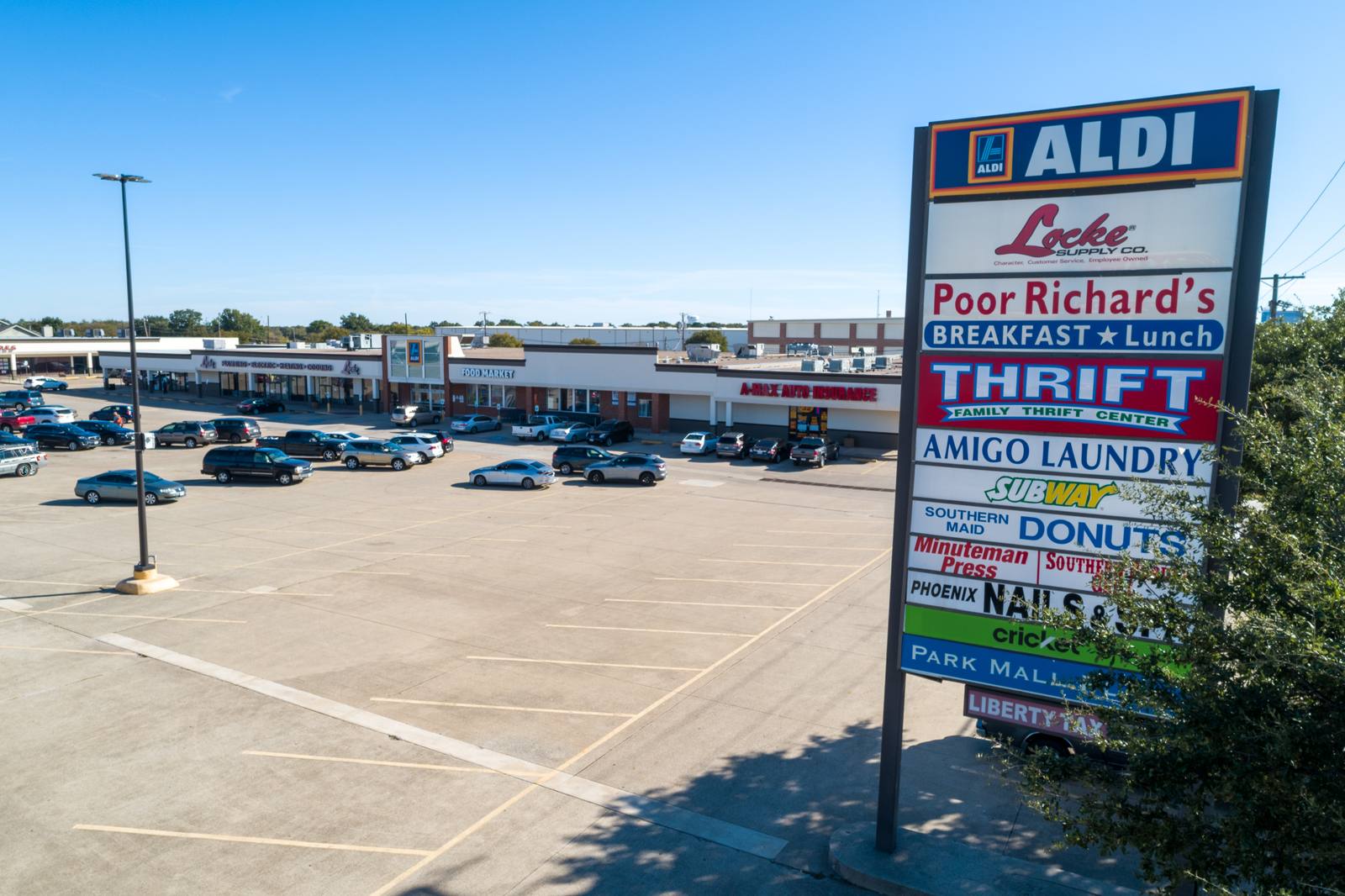 A tall, multi-sign commercial sign stands in a paved parking lot filled with cars, with a row of storefronts visible in the background under a clear blue sky.