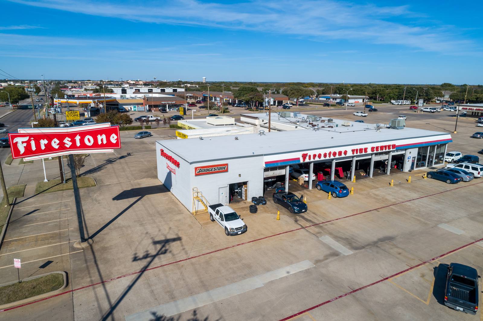 Aerial view of a Firestone Tire Center with multiple cars, including a blue sedan and a white pickup, parked in its lot under a clear blue sky.