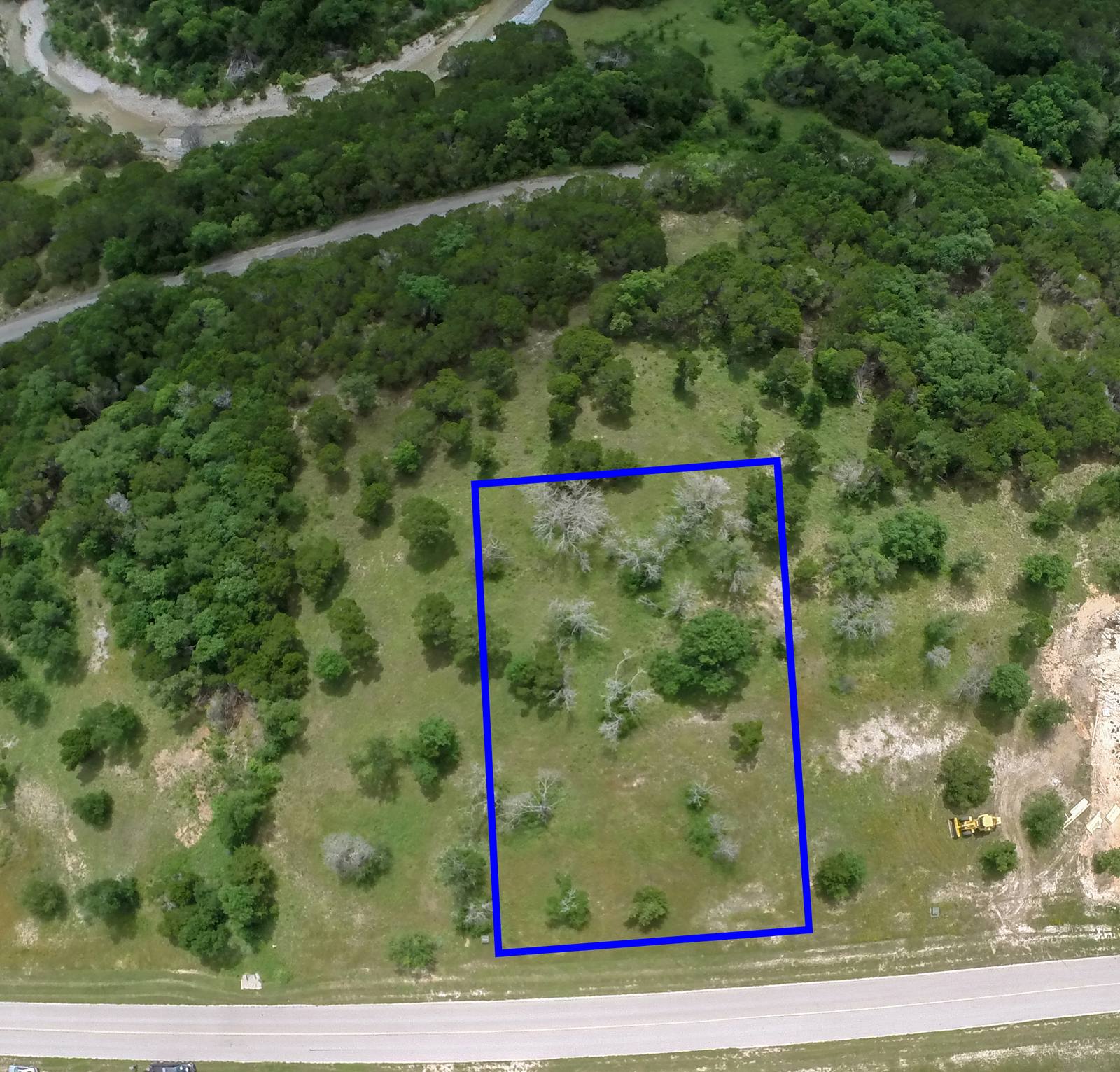 Aerial view shows a blue-outlined rectangular plot within a wooded, grassy landscape, bordered by a road and adjacent to construction equipment.