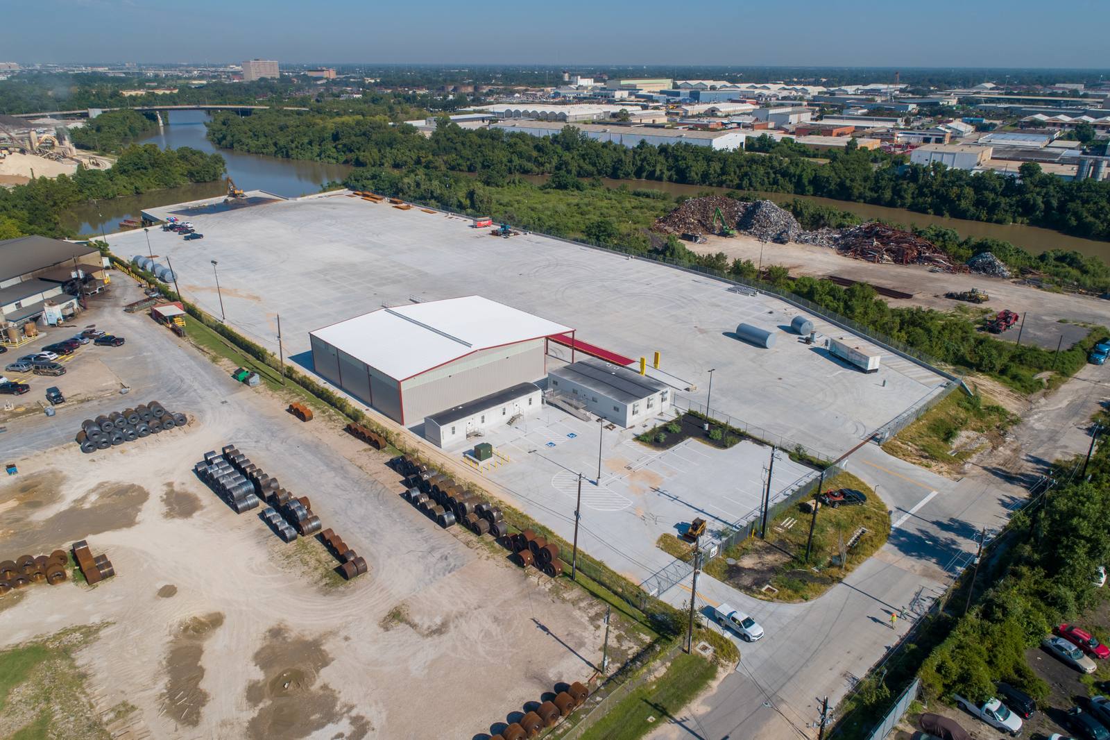 Aerial view of a large industrial complex with a white-roofed building, surrounded by paved lots, stacked metal coils, and a river with a bridge in the distance.