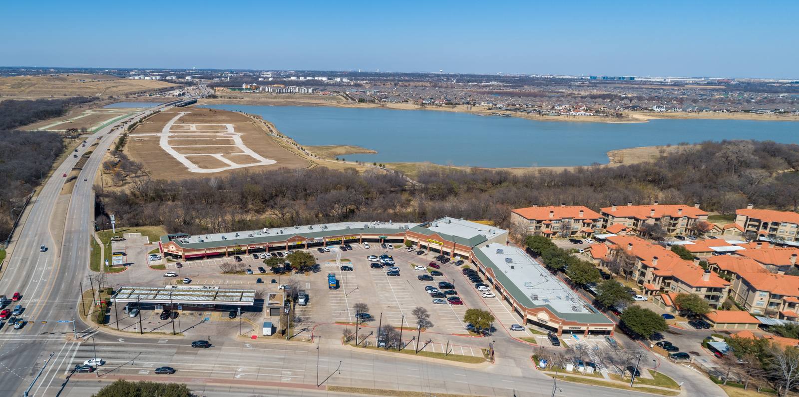 Aerial view shows a commercial complex with parking lots, adjacent residential buildings with terracotta roofs, and a large lake with a white geometric patterned shoreline.