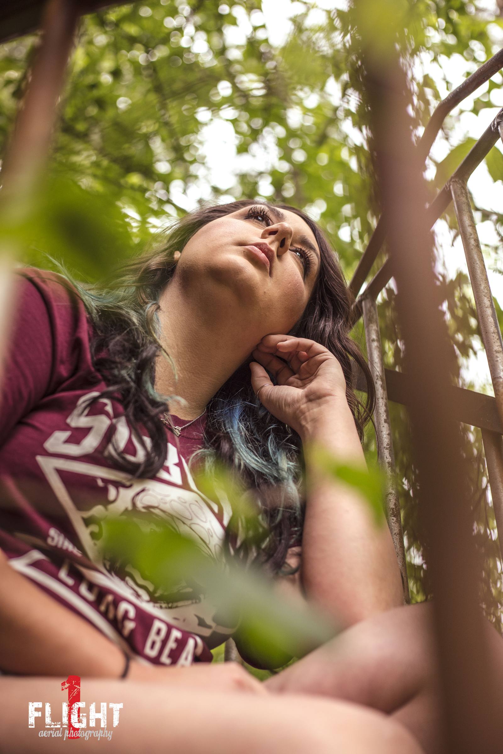 A low-angle portrait of one woman with dark hair and blue highlights, wearing a maroon graphic tee, resting her chin on her hand amidst green foliage and a metal railing.