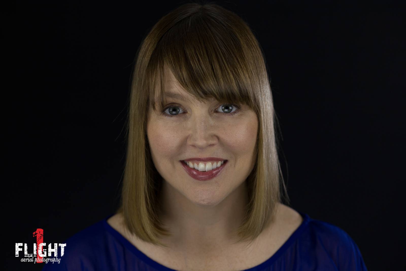 Head-and-shoulders portrait of a smiling woman with shoulder-length brown hair and bangs, wearing a blue top, against a plain black background.