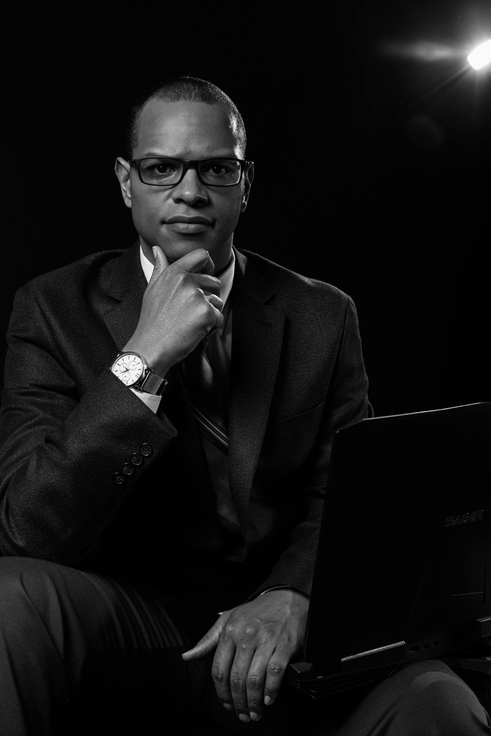 Black-and-white studio portrait of one man in a suit and glasses, seated with a laptop, hand to chin, against a dark background with a bright light flare.
