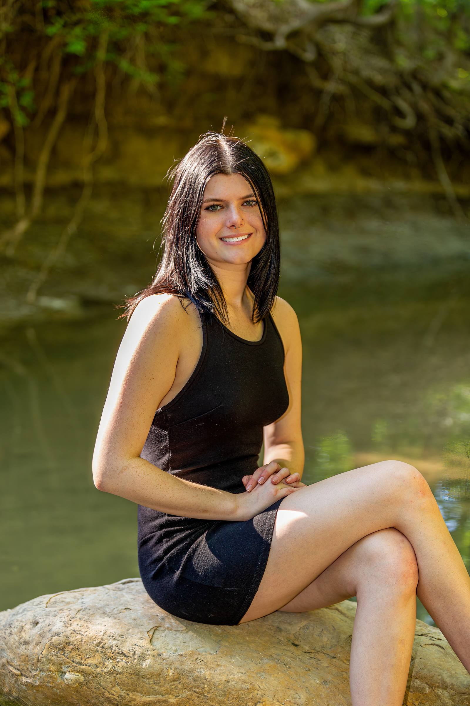 A woman in a black dress sits on a rock by water, smiling at the camera in a wider portrait with blurred foliage and trees in the background.