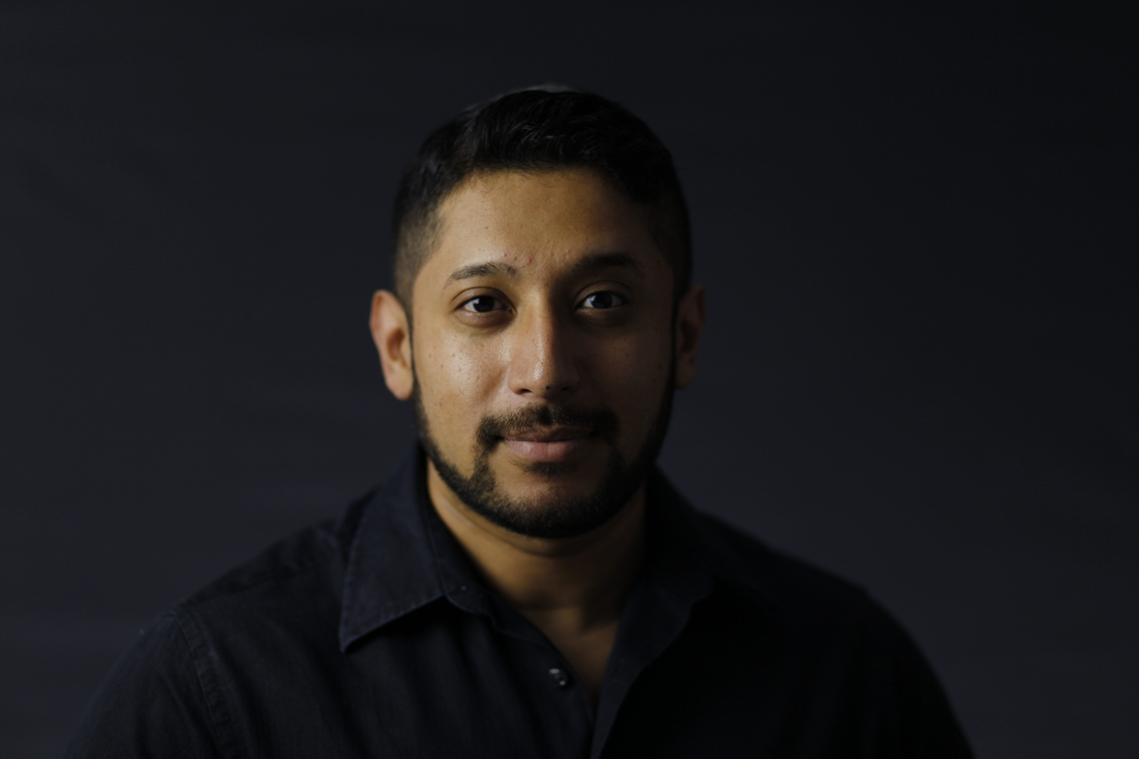 Head-and-shoulders portrait of a man with short dark hair and beard, wearing a dark collared shirt, against a plain dark background.