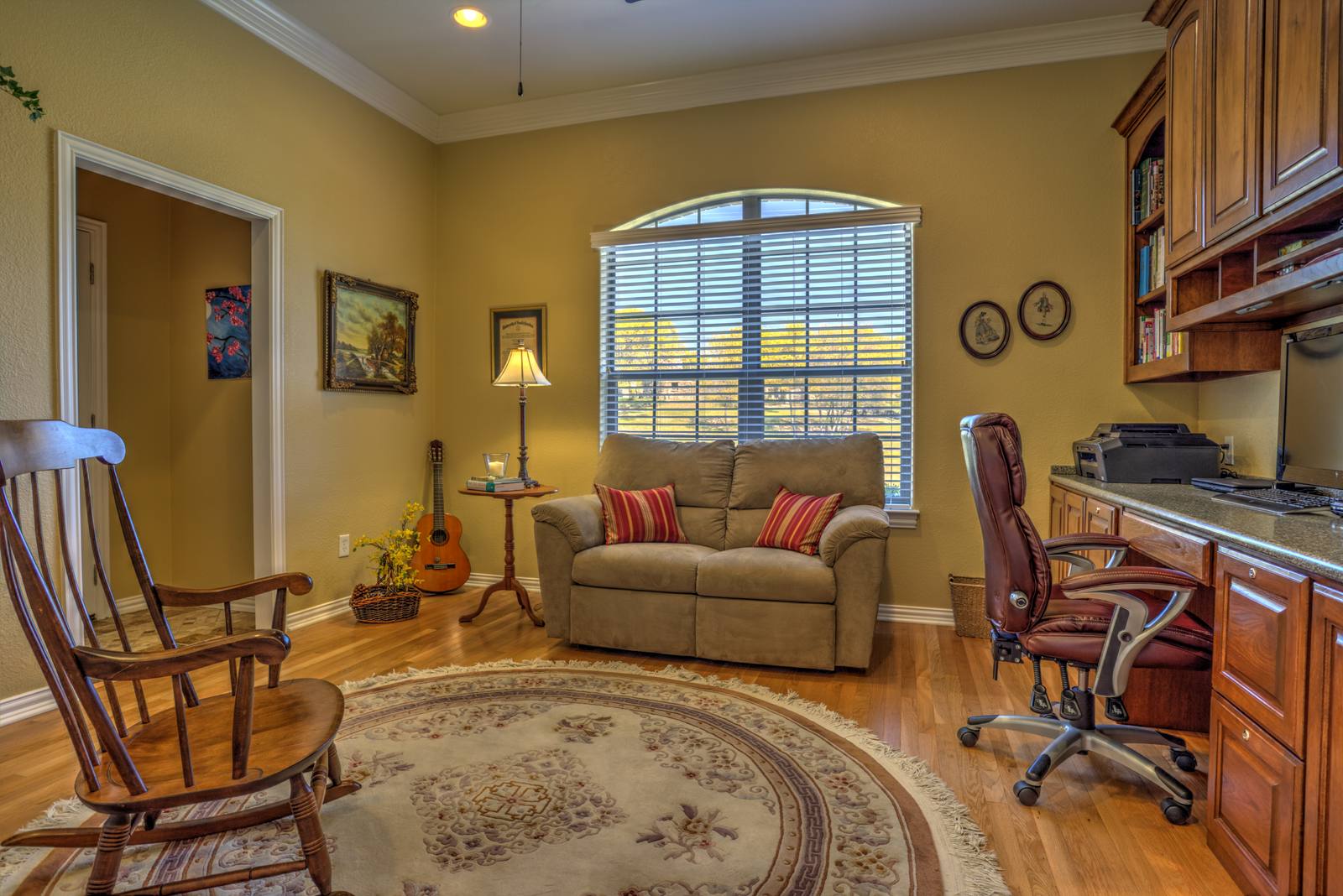 A furnished home office and living area with a rocking chair, sofa, desk, guitar, and window with blinds, no people visible.