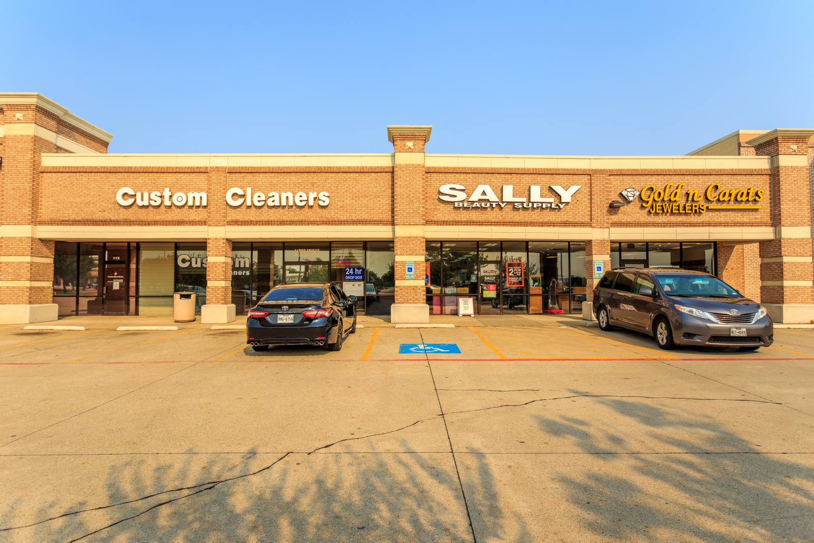 Three storefronts with visible signage: "Custom Cleaners," "SALLY BEAUTY SUPPLY," and "Golden Carats JEWELERS," each with parked cars in front under clear sky.